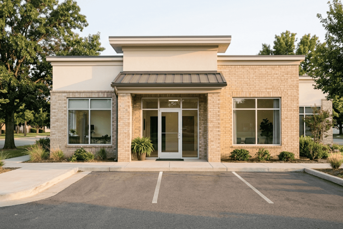 Front view of a small brick office building with glass double doors and large windows, parking lot at the front.